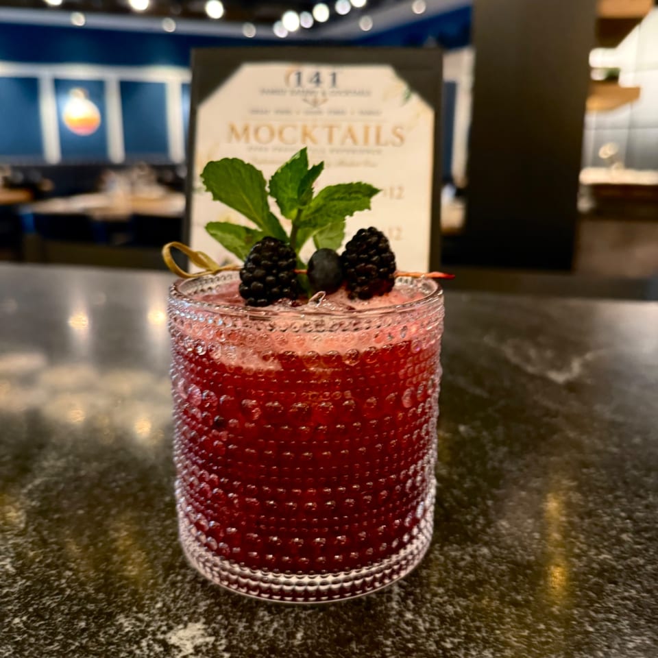 Red berry mocktail in a textured glass, garnished with blackberries, mint leaves, and a red straw on a restaurant bar.