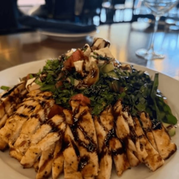 Grilled chicken with balsamic glaze, fresh greens, tomatoes, and mozzarella on a white plate.